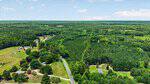 Aerial view of a rural landscape featuring Tbd Hicksboro Road winding through scattered houses, green fields, and dense forests under a partly cloudy sky near Henderson.