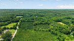 Aerial view of a lush, green forest landscape near Tbd Dabney Road in Henderson, with scattered houses, open fields, and a road running along the left side under a partly cloudy sky.