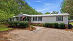 Single-story gray manufactured home with white trim, black shutters, and a metal roof at 104 Pineview Drive, Louisburg. An American flag hangs by the front porch. The yard has green bushes, trees, and a gravel driveway under a blue sky.