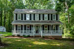 A two-story house at 1011 Homestead Place in Zebulon features gray siding, white trim, black shutters, and a wide front porch with white railings, all surrounded by trees and grass.