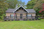 Single-story tan house with black trim, front porch, and gabled roof at 33 American Legion Road, Franklinton. An American flag hangs near the steps. The yard is grassy with some shrubs and large trees surrounding the house.