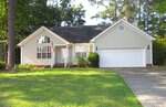 Single-story light beige house with two-car garage, gabled roof, arched front window, small porch, and American flag. Located at 2723 Clifton Avenue in Creedmoor, this charming home is surrounded by a green lawn and tall trees, with a paved driveway in front.
