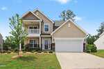 Charming two-story beige house at 90 Bourne Drive, Franklinton, featuring a welcoming front porch, balcony, white trim, and a spacious two-car garage. A small tree and well-maintained shrubs adorn the front yard, with a driveway that leads directly up to the garage.