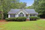 A small white house with dark shingles at 140 Fairway Lane, Zebulon features a central arched window, front steps, bushes, and a grassy yard, all surrounded by tall trees.
