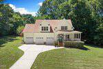 A two-story house at 1084 Whip Lane, Stem, featuring a charming front porch, three-car garage, and large driveway, is surrounded by green grass and trees under a blue sky with scattered clouds.