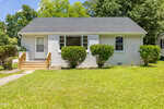 Charming small white house with a dark roof at 605 E Montgomery Street, Henderson, featuring a central front window, wooden steps leading to the door on the left, and neatly trimmed bushes and green lawn in front. Trees and blue sky provide a picturesque background.