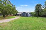 A blue single-story house with a gray roof sits on a spacious grassy yard at 962 Laurel Mill-Centerville Road, Louisburg, surrounded by trees and a dirt driveway, under a clear blue sky.