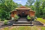 A wooden cabin with a covered front porch sits surrounded by lush green trees on 3686 Sandy Creek Road in Franklinton. Steps lead to the entrance, flanked by potted plants and landscaped garden beds. The sky is clear and blue.
