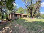 Single-story brick house with a sloped roof, large windows, and a grassy front yard surrounded by tall trees under a blue sky with scattered clouds, ideally situated at 2560 Mangum Avenue in tranquil Creedmoor.