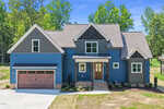 Modern two-story blue house with gabled roof, brick accents, brown double garage door, central front steps, and landscaped front yard on 270 Pilot Ridge Road in Zebulon, surrounded by trees and blue sky in the background.