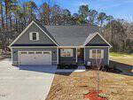A modern gray house with white trim at 12867 Mallard Rock, Zebulon, featuring a two-car garage, front porch, and a wide concrete driveway. The yard is partly grassed with a small tree and bordered by woods in the background under a clear sky.