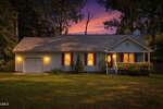A single-story house with a front porch, garage, and red shutters is warmly lit at dusk, surrounded by trees and a lawn, under a colorful sunset sky.