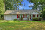 Single-story light blue house with red shutters, a front porch, attached garage, and a green lawn, surrounded by tall trees under a clear blue sky.