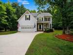 White two-story house with attached garage at 105 Highview Drive, Youngsville, featuring an American flag, wide driveway, manicured lawn, bushes, and trees on both sides. Set against a backdrop of tall green trees under a blue sky with scattered clouds.