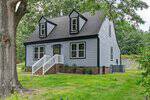 A two-story gray house with black roof, white trim, brick foundation, and two dormer windows sits gracefully at 110 Quail Ridge in Henderson. There are white railings on the front steps, green grass, trees, and a fenced yard in the background.