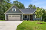 Single-story gray house with white trim at 12867 Mallard Rock, Zebulon, featuring a two-car garage, front porch, and gabled roof. This charming home is surrounded by green grass, a small tree, and wooded trees in the background under a blue sky.