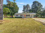 Single-story white house with a gray roof, attached carport, large front yard, trees, and a dirt driveway under a clear blue sky in Henderson at 1020 Jackson Royster Road.