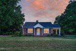 A small brick house with white trim and blue shutters is lit from within at sunset on 148 Baptist Church Road, Henderson. The lawn is green with shrubs, and tall trees frame the home on both sides.