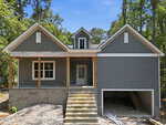 Gray house under construction at 102 Mayan Drive, Louisburg, with a front porch, steps leading to the main door, and an unfinished garage underneath. Surrounded by trees, the house features white trim and an upper dormer window.