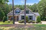 A modern two-story house with gray siding, stone accents, and a front porch, 105 Carriden Drive in Youngsville is surrounded by neatly trimmed bushes, trees, and a wooded background. A paved driveway leads to a side garage.