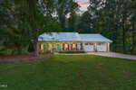 A single-story house with a metal roof, front porch, and attached garage sits surrounded by trees and grass at dusk on 137 Sacred Fire Road in Louisburg. An American flag hangs by the front door.