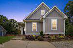 A modern gray house with white trim, tall windows, and a brick foundation sits at 408 S Main Street in Franklinton, set against a blue and purple evening sky. Shrubs and small bushes line the front yard near a concrete driveway.