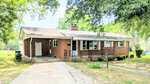 Charming single-story brick house at 103 Hayes Street, Louisburg, featuring a welcoming front porch, white trim, and a classic chimney. A driveway leads to a convenient side entrance. The yard offers lush bushes and mature trees, with the home nestled under partial shade for added comfort.