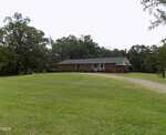 A single-story brick house with a green roof sits on a large grassy lawn at 3600 NC 561 Hwy, Louisburg, surrounded by trees. A gravel driveway curves toward the right side of the house under an overcast sky.