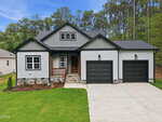 A modern single-story house at 117 Deer Cross Drive in Louisburg features gray siding, black trim, two black garage doors, a front porch with wooden railing, stone accents, a concrete driveway, green lawn, and trees in the background under a blue sky.