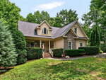 A tan house with stone accents, gabled roof, front porch, and columns sits gracefully at 1514 Anterra Drive in Wake Forest, surrounded by green trees, bushes, and a grassy lawn under a blue sky.