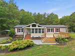 A single-story house at 7569 NC 96, Oxford, features beige siding, red shutters, a front porch with steps and a ramp, landscaped bushes, and trees in the background under a clear blue sky.