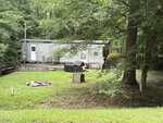 A small, light gray mobile home sits among trees at 108 Natchez Drive, Louisburg, featuring a wooden porch, grill, and green lawn in front. A few outdoor items and shaded areas are visible around the yard.