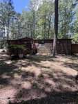 A rustic, single-story wooden cabin with a red deck sits in a sunlit, wooded area on 197 E Holly Lane, Henderson, surrounded by tall trees and patches of grass.