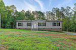 A single-story manufactured home with light siding and dark shutters stands on a grassy lawn at 3952 Hicksboro Road in Henderson, surrounded by trees. A small front porch with white railings leads to the entrance.