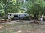 A white Sportsmen RV is parked in a wooded campsite at 126 Geronimo Drive, Louisburg, featuring a wooden deck, picnic table, small fire pit, and signs. Trees surround the area, providing shade and greenery.
