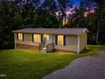 A single-story house with lit windows, beige siding, brick foundation, and a small front porch sits on a grassy lawn bordered by trees at sunset at 100 Rough Lane, Louisburg. A gravel driveway leads up to the house.