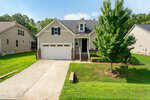 A beige single-family house at 165 Olde Liberty Drive in Youngsville features a two-car garage, brick accents, front porch, green lawn, small tree, and a concrete driveway, set between two similar houses on a sunny day.