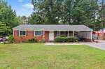 Charming single-story red brick house with a gray roof, white trim, and a carport on the right, located at 1277 David Avenue in Henderson. There are shrubs in front and a large front yard with a concrete path, surrounded by trees and greenery under a blue sky.