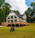 A beige two-story house with large windows, a black roof, and a wraparound wooden deck sits on a grassy lawn bordered by tall trees under a partly cloudy sky at 89 Friendly Lane in Henderson.