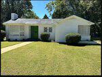 A single-story white brick house with a green front door, large windows, and well-kept bushes sits at 203 W Rockspring Street in Henderson. The lawn is neatly mowed, and trees are visible in the background under a clear sky.