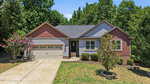 Charming single-story house at 265 N Ridge Drive, Louisburg, featuring beige and red siding, a two-car garage, a gray shingled roof, shrubs, a small tree in front, and surrounded by green trees under a clear sky.