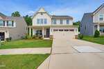 Two-story suburban house with beige siding, a covered front porch, two-car garage, and a wide driveway located at 1165 Spring Meadow Way in Wake Forest. The yard features green grass and minimal landscaping. Other similar homes are visible on each side.