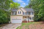 Charming two-story beige house with black shutters, white trim, a front porch, and a two-car garage located at 108 Cedar Street in Butner. Surrounded by trees, the house sits on a sloped lot with a concrete driveway and steps leading to the entrance.
