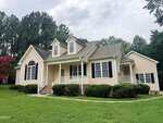 A beige, single-story house at 7581 NC 96 in Franklinton features a welcoming front porch, black shutters, dormer windows, and neatly trimmed shrubs. It sits on a green lawn with trees in the background under a cloudy sky.