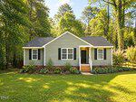 A small, single-story house at 108 Bristol Court in Youngsville features gray siding, white trim, black shutters, and a dark front door. Surrounded by a green lawn, flower beds, and tall trees in the background under a blue sky, this charming home offers both comfort and curb appeal.
