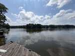A wooden dock extends over a calm lake at 126 Big Horn Drive, Louisburg, reflecting trees and a partly cloudy sky, with forested shoreline visible in the distance.