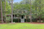 A two-story beige house at 3584 Creekstone Way, Wake Forest, featuring a wide front porch with white railings and steps, is surrounded by tall trees and greenery, with a well-maintained lawn in the foreground.