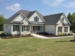 A modern single-story house with white siding, stone accents, dark shutters, and a gray roof sits at 125 Water Willow Lane in Zebulon. It is surrounded by green grass, mulched flower beds, and trees in the background under a blue sky.
