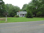 A small gray house with white trim sits back from a two-lane road at 1121 S Cokesbury Road in Henderson, surrounded by green grass, trees, and shrubs on a cloudy day. A mailbox is visible near the driveway on the left.