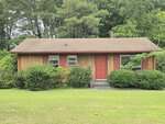 A small, single-story wooden house with a red door, red shutters, and a brown roof sits at 140 Evans Road in Henderson, surrounded by green bushes and trees, with a well-kept lawn in front.